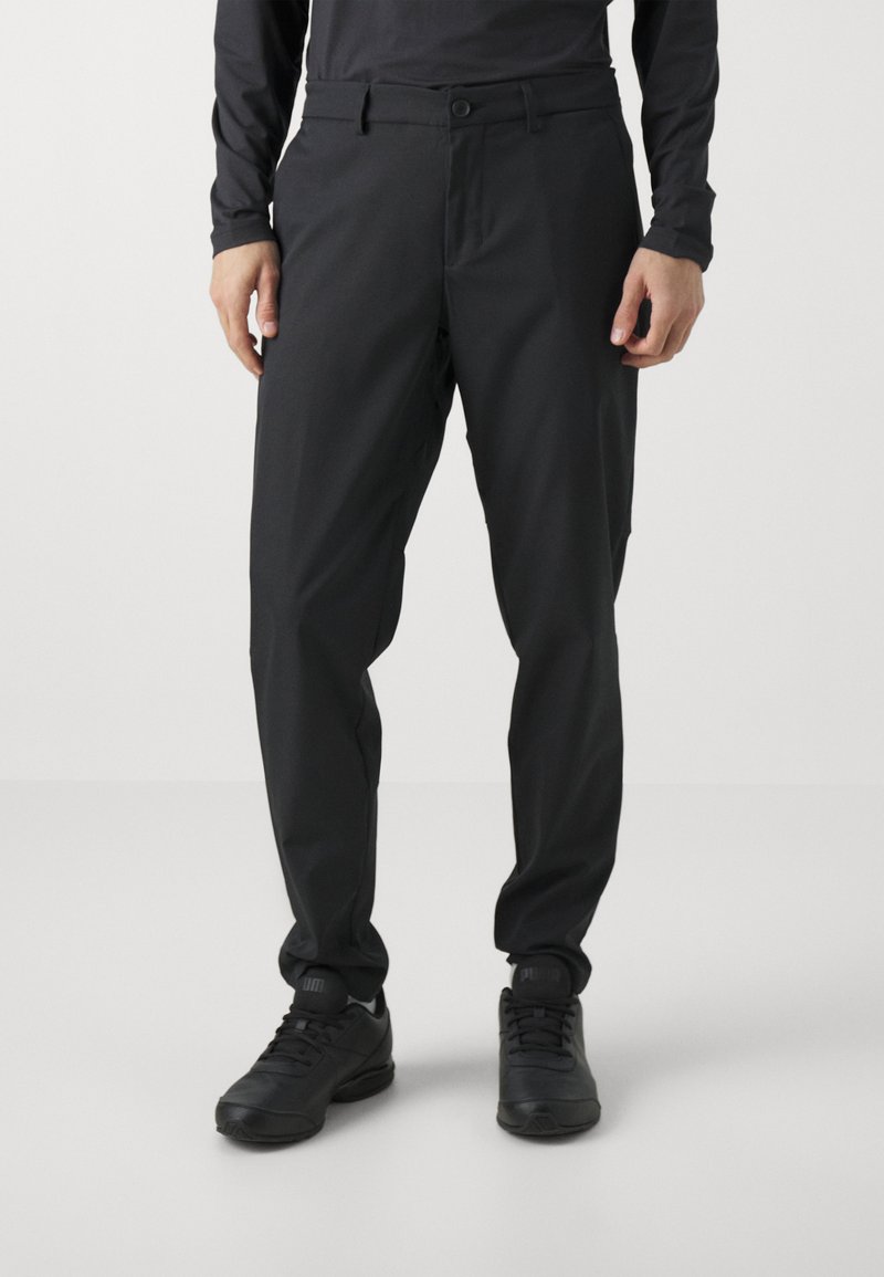 Black tapered trousers made from smooth, lightweight fabric. Features a belt loop waistband, front pockets, and ankle cuffs. Paired with black sneakers.