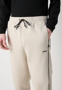 Beige sweatpants med snoretræk i taljen, sidelommer med lynlås og sorte detaljer. Har et logo på venstre lår og en glat tekstur.