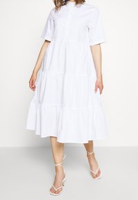 Abito bianco con bottoni, maniche corte, design a livelli e orlo fluido. Indossato con sandali bianchi con cinturini. Tessuto di cotone liscio.