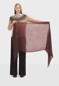 Écharpe bordeaux transparente tenue à l'extérieur, avec une texture légère et des bords effilochés. Portée avec un pull à motif beige et un pantalon noir.