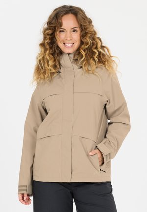 Frau mit lockigem Haar, die eine beige Jacke und schwarze Hosen trägt, lächelt mit einer Hand in der Jackentasche vor schlichtem Hintergrund.