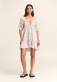 Gestreepte jurk met een diepe halslijn, pofmouwen en een gerufflede zoom. De stof heeft multicolor verticale strepen. Strik aan de hals.