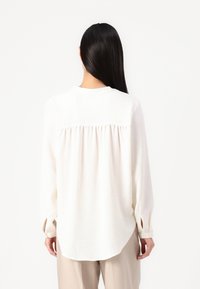 Weiße Bluse mit langen Ärmeln, leicht strukturiertem Rücken, gerafftem Detail an den Schultern und abgerundetem Saum. Sichtbare Knopfmanschetten.