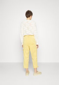 Een persoon staat met de rug naar een muur, gekleed in gele cropped broeken, beige schoenen en een transparante witte blouse met lange mouwen en bloemenpatronen.