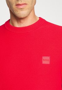 Suéter rojo texturizado con escote redondo y un sutil parche de logo cuadrado etiquetado como "BOSS" en la zona del pecho izquierdo.