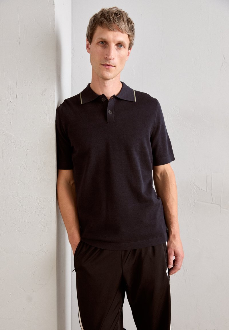 Polo shirt sombre avec manches courtes et col contrasté. Texture lisse, fermeture à deux boutons. Associé à un pantalon noir avec des accents sur le côté.