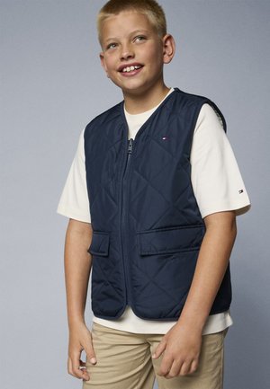 Gilet matelassé marine avec deux poches avant, fermeture éclair, et un logo contrasté sur la poitrine, porté par-dessus un t-shirt crème et un pantalon beige.