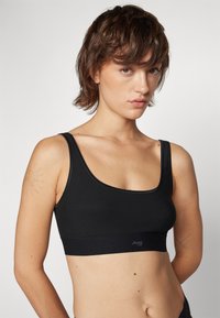 Soutien-gorge de sport noir en tissu doux avec un col en scoop et de fines bretelles. Présente une large bande élastique avec un détail de logo en bas.