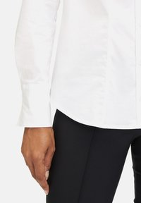 Rechte Hand einer Person, die ein weißes Langarmshirt und schwarze, eng anliegende Hosen trägt, vor einem schlichten weißen Hintergrund.