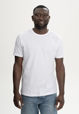 Lachende man met kort haar, gekleed in een effen wit T-shirt met korte mouwen en een blauwe spijkerbroek, staand tegen een lichte achtergrond.