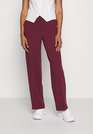 Kobieta nosząca białe trampki, luźne bordowe spodnie i biały bezrękawnik z ażurowymi detalami.