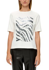 Vit t-shirt med kort ärm som har silver- och svarta paljettvågformer. T-shirten har en avslappnad passform och är kombinerad med svarta läderbyxor.