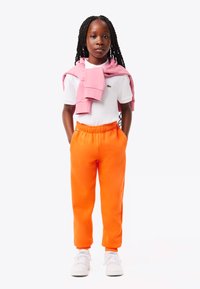 Kind met een wit T-shirt met korte mouwen, oranje trainingsbroek en een roze trui om de schouders; witte sneakers maken de look compleet.