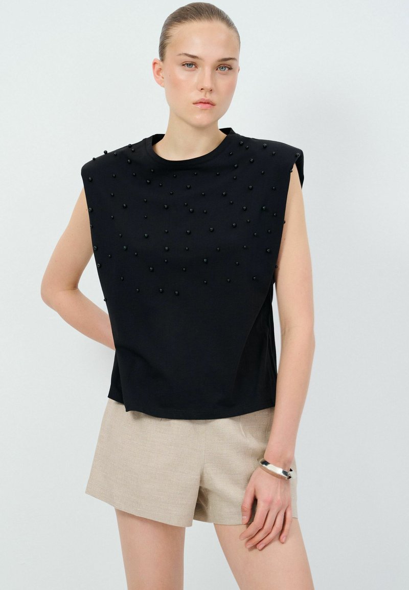 Top senza maniche nero con perline nere in rilievo sul fronte, dal fit largo e con scollatura rotonda. Abbinato a pantaloni corti beige.