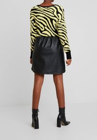 Žena oblečená v yellow and black zebra print svetru, černé kožené mini sukni a černých vysokých kotníkových botách stojí na pozadí bez vzoru.