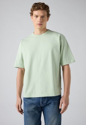 Νέος άνδρας με σγουρά μαλλιά, που φοράει μια απλή ανοιχτοπράσινη κοντομάνικη μπλούζα και μπλε τζιν, στέκεται μπροστά σε ένα απλό γκρι φόντο.