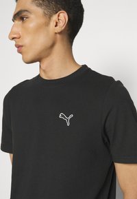 Sort t-shirt lavet af bomuld med rund hals. Har et hvidt broderet Puma-logo på venstre bryst. Korte ærmer, glat tekstur.