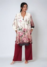 Floral-print long shirt in cream, featuring pink and green flowers, a collared neckline, and balloon sleeves. Worn over burgundy wide-leg pants.