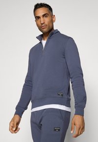 Navy blue zip-up sweatshirt in soft fabric with long sleeves, high collar, and logo tag at the hem. Worn over a white top and matching joggers.