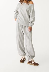 Lichtgrijze sweatshirt met off-shoulder ontwerp, gecombineerd met bijpassende grijze joggingbroek met elastische boorden en bruine sneakers met crèmekleurige accenten.