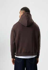 Bruine hoodie met een grote capuchon, gestructureerde zijkanten en een ontspannen pasvorm. Gecombineerd met donkere spijkershorts. Eronder een effen wit T-shirt.