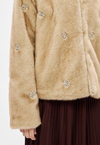 Casaco de pele falsa cor castanha clara com aplicações de joias. Mangas compridas e textura macia. Usado por cima de uma saia plissada castanha.