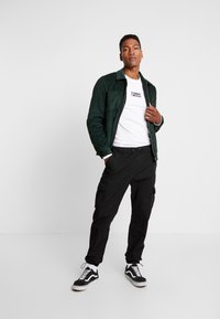 Jachetă verde din catifea, purtată peste un tricou alb cu logo, asortată cu pantaloni cargo negri și adidași negri cu dungi albe.