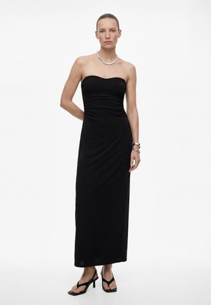 Kvinde iført en sort stropløs maxikjole, sorte sandaler med hæl, sølvhalskæde og armbånd, stående mod en hvid baggrund.