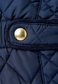 Tejido acolchado azul marino con un botón dorado de la marca "Joules" y costuras texturizadas. Presenta una superficie lisa y un acabado suave.