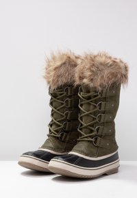 Ein Paar grüne Winterstiefel mit schwarzen Gummisohlen, Schnürungen vorne und braunem Kunstfellbesatz am oberen Rand.