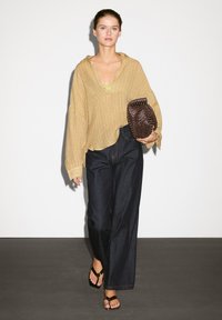 Beige oversized pletený svetr s výstřihem do V, spojený s širokými tmavohnědými džínami a černými sandály. Drží tkanou tmavě hnědou kabelku.