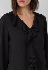 Zwarte blouse met een v-halslijn en ruches, lange mouwen en een soepele, gestructureerde stof.