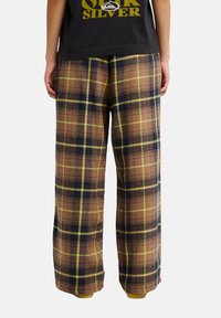 Personne portant un pantalon large à carreaux marron, noir et jaune et un t-shirt noir Quiksilver, vue de dos sur fond blanc.