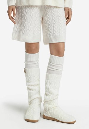 Weiße Strickshorts mit geripptem Saum, kombiniert mit Zopfmuster-Beinwärmern und weichen, weißen Schuhen mit strukturierter Oberfläche und Gummisohle.