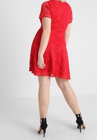 Röd spetsklänning med korta ärmar, utsvängd kjol, blommönster och en åtsittande bodice, kombinerad med svarta högklackade sandaler med remmar.