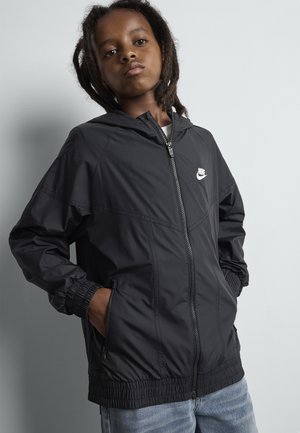 Bambino con dreadlocks che indossa una giacca nera con zip Nike e jeans blu, in piedi contro una parete chiara e uniforme con le mani in tasca.