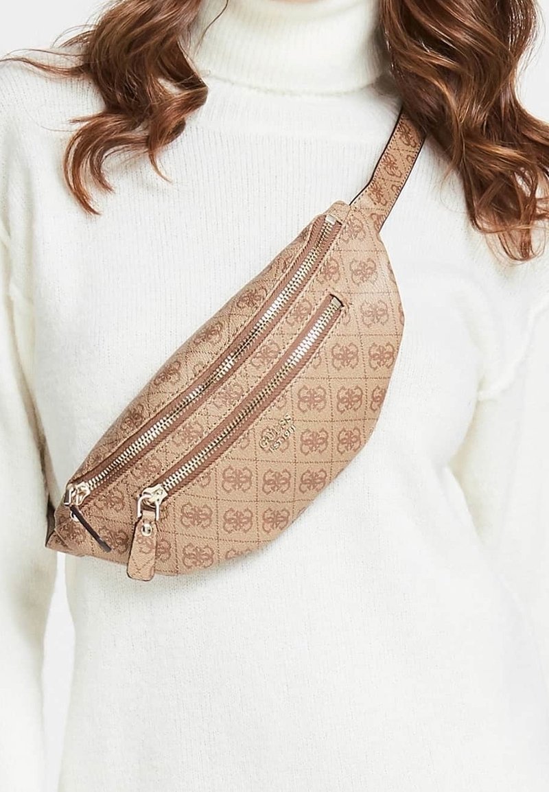 Femme portant un sac bandoulière beige à motifs avec deux fermetures éclair argentées sur un pull à col roulé blanc.