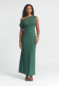 Zelené šaty na jedno rameno s drapovaným materiálem, užším střihem a délkou až na podlahu. Disponují hladkým povrchem a jemným leskem. Černé sandály na tenkých páscích.
