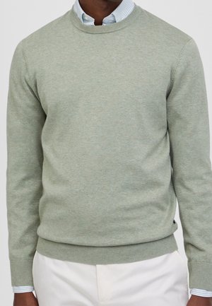 Mann trägt hellgrünen Rundhalspullover über einem blau-weiß gestreiften Hemd mit Kragen und weißen Hosen.