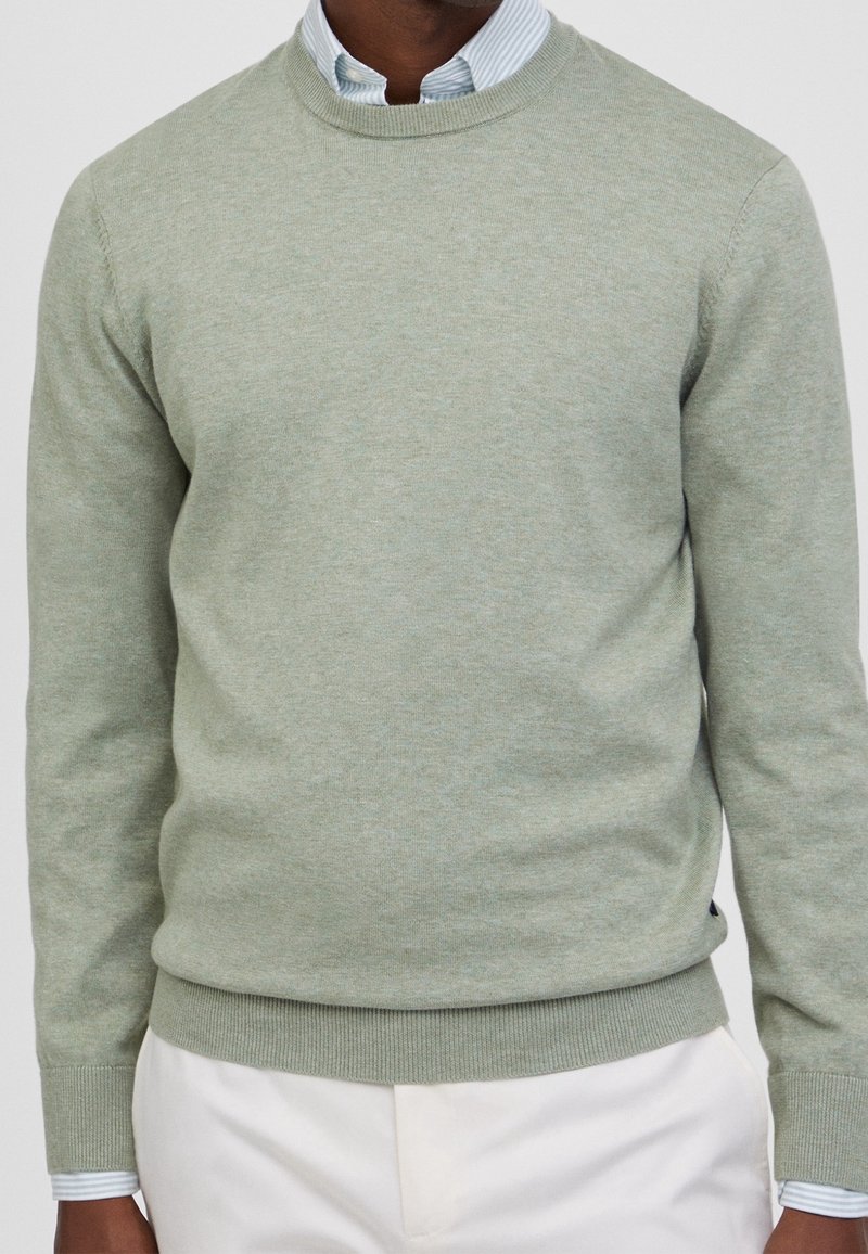 Homme portant un pull léger vert clair à col rond sur une chemise rayée bleue et blanche à col avec un pantalon blanc.