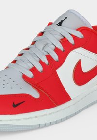 Παπούτσι τύπου sneaker χαμηλό με λευκά και κόκκινα δερμάτινα πάνελ, λευκά κορδόνια, μαύρο λογότυπο Nike Swoosh στη μύτη, κόκκινο Swoosh στο πλάι και λογότυπο Jumpman στη γλώσσα.