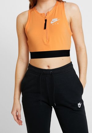 Vrouw draagt een oranje mouwloos Nike Air crop top met rits aan de voorkant en zwarte Nike joggingbroek met trekkoord en zakken.