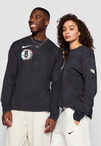 Crne majice dugih rukava s okruglim izrezom i rebrastim manžetama. Lijeva majica sadrži šareni logo Brooklyn Netsa i Nike swoosh.