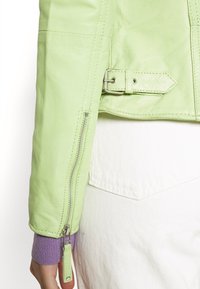 Manche de veste en cuir vert clair avec fermeture éclair et détail de boucle argentés, portée sur un poignet violet et un pantalon blanc.