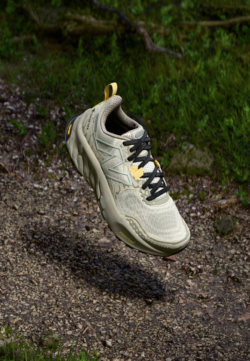 Lys beige treningssko med mesh-overdel, svarte lisser, gummisåle og gule detaljer. Har en tydelig logo på siden og strukturerte detaljer.