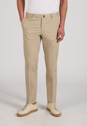 Mężczyzna w beżowych obcisłych spodniach, białych niskich trampkach i białej koszulce wsuniętej w spodnie, stoi z jedną ręką w kieszeni.