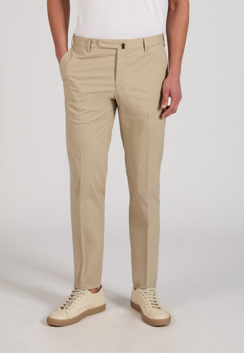 Homme portant un pantalon beige slim, des baskets blanches basses, et un t-shirt blanc rentré, debout avec une main dans la poche.