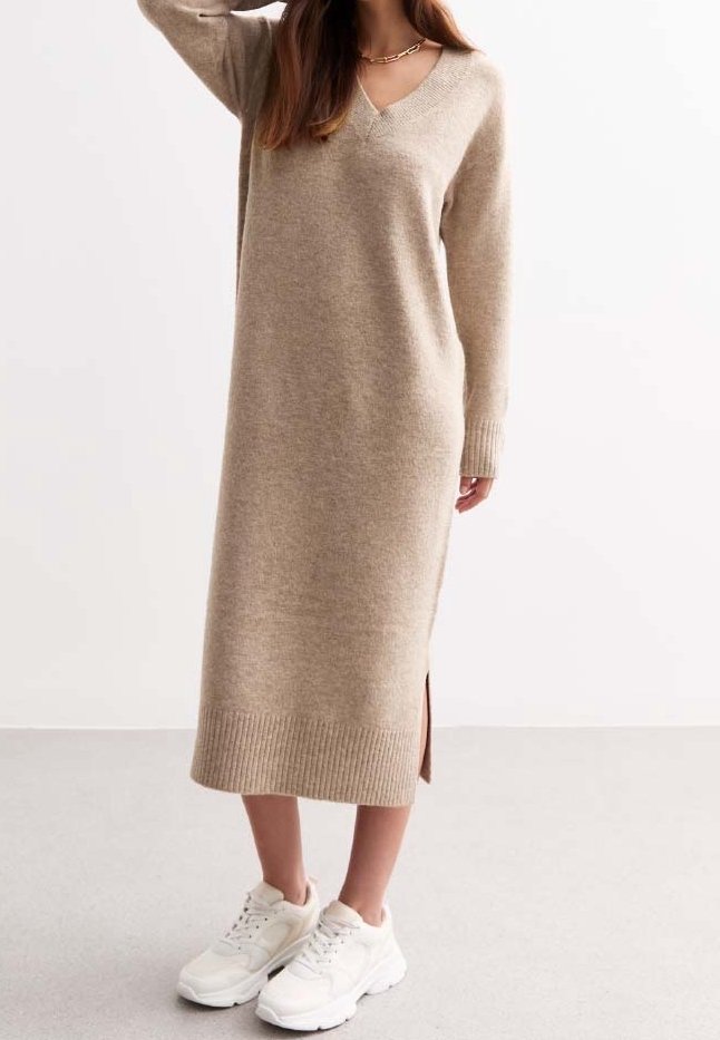 Femme portant une robe pull longue en maille beige avec une fente sur le côté, des baskets blanches épaisses et un collier chaîne en or, debout devant un fond uni.