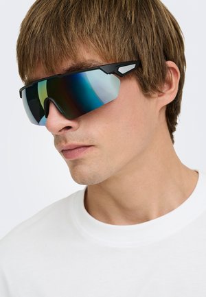 Jeune homme aux cheveux châtain clair portant de grandes lunettes de sport noires à verre miroir et un t-shirt blanc uni à col rond.