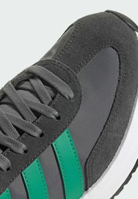 Gros plan sur une sneaker grise avec des rayures latérales vertes, en suède texturé, tige en textile, lacets gris et détail de semelle blanche.